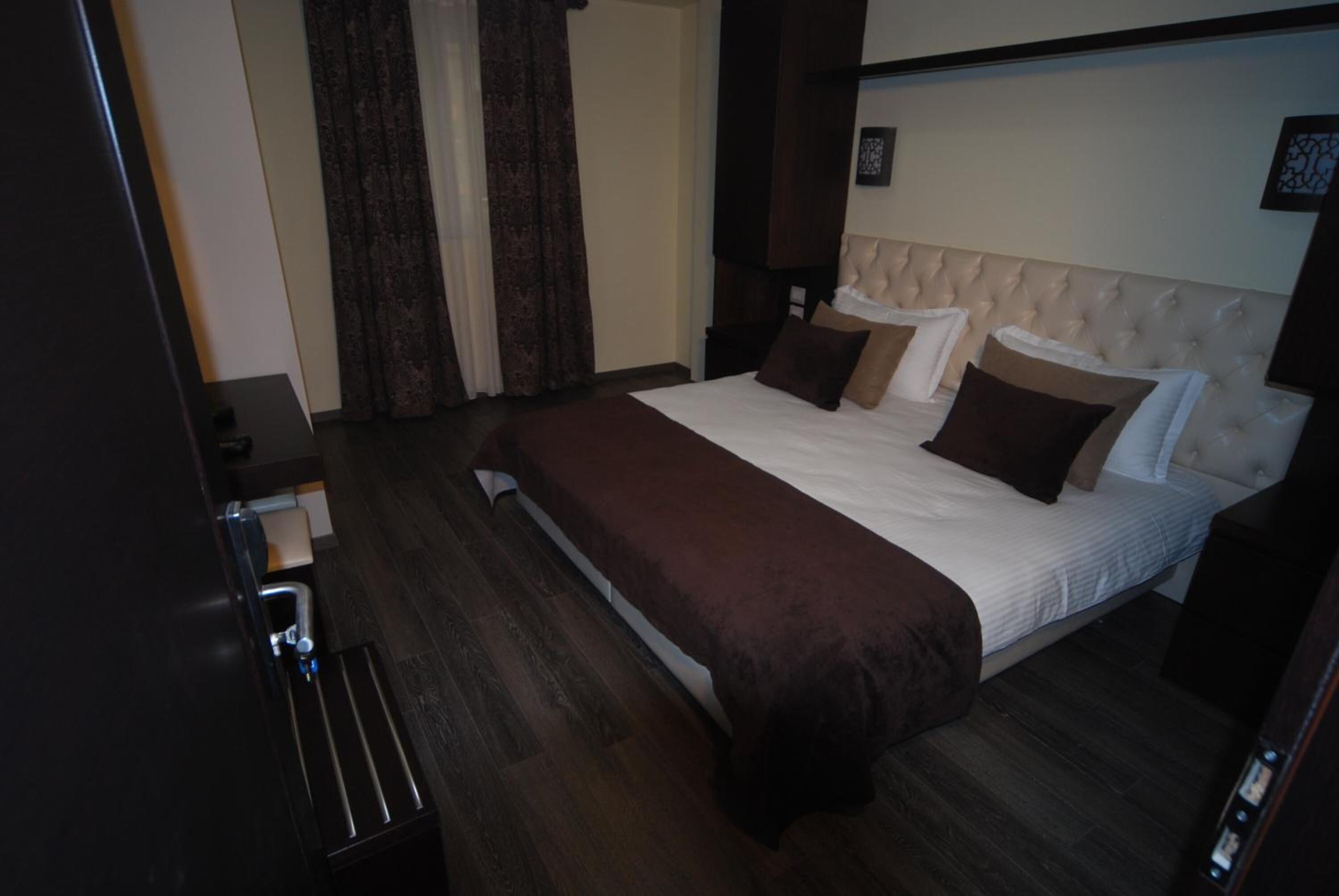 A hotel room with a large bed, dark wood flooring, a dark brown bedspread, and a white headboard.