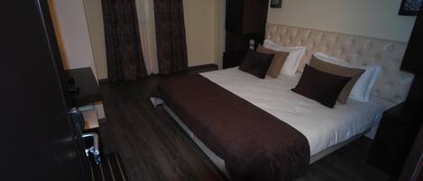 A hotel room with a large bed, dark wood flooring, a dark brown bedspread, and a white headboard.