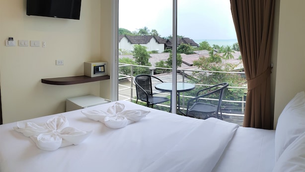 A hotel room with a bed, a TV mounted on the wall, a small table with chairs, and a balcony with a view of the sea and buildings. - Top Hostel Samui (Koh Samui)