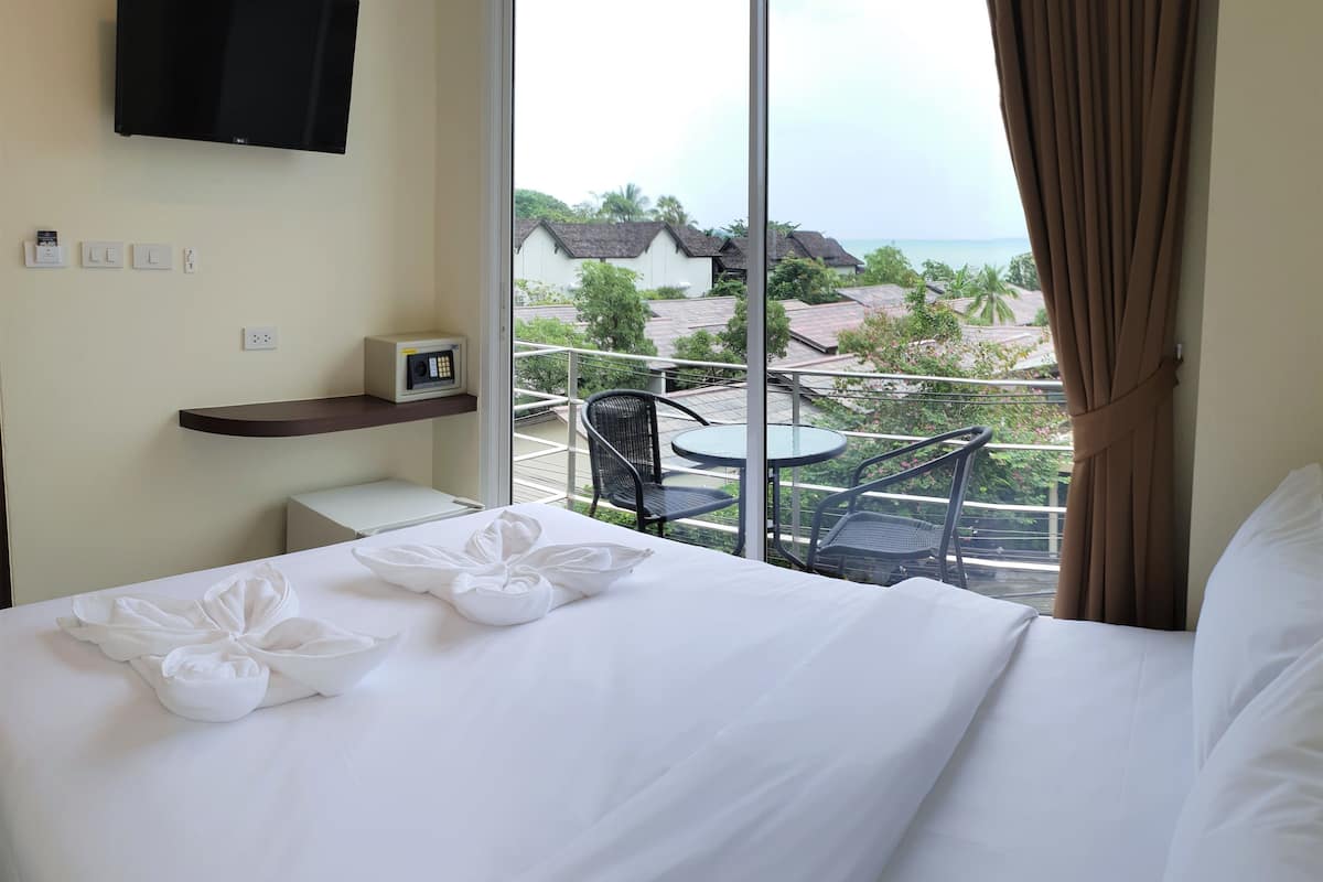 A hotel room with a bed, a TV mounted on the wall, a small table with chairs, and a balcony with a view of the sea and buildings.