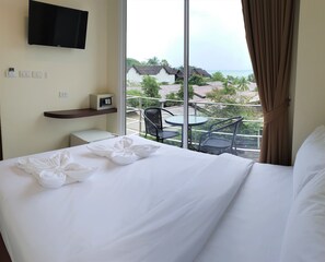 Quarto de hotel com uma cama, televisão fixada na parede, uma pequena mesa com cadeiras e varanda com vista para o mar e edifícios.