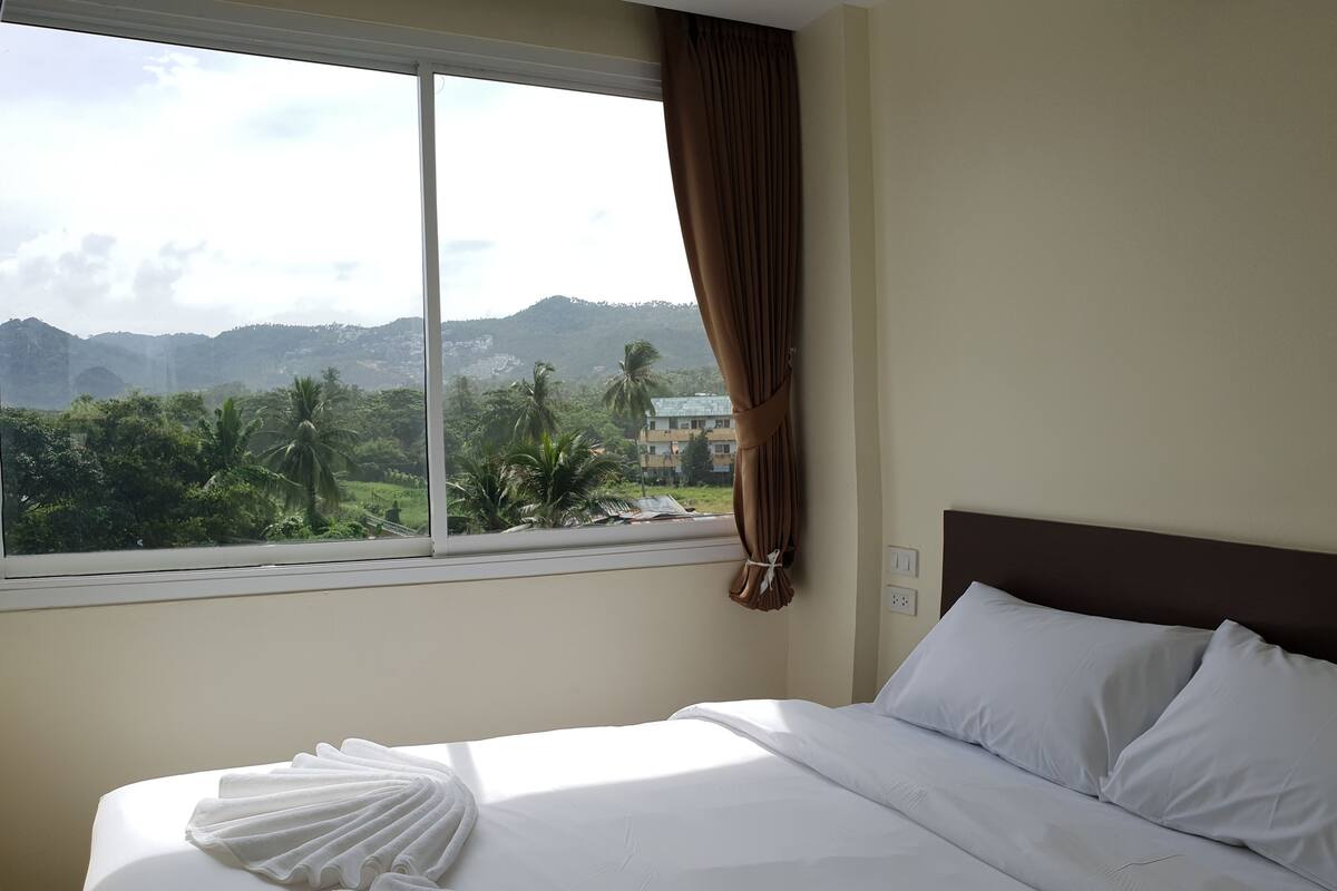 A hotel room with a large bed, a view of greenery and hills, and a window with curtains.
