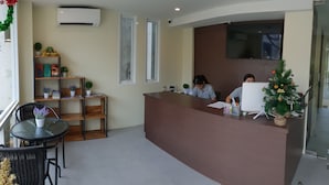 A reception area with a brown counter, two windows, a small Christmas tree, and a bookshelf with plants. - Top Hostel Samui (Koh Samui)