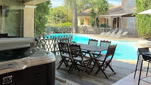 Un patio exterior con un jacuzzi, mesa de comedor y sillas, con vistas a una piscina y un jardín.