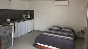 A compact room with a kitchenette, a bed with patterned bedding, and a ceiling-mounted air conditioning unit. - House with independent studio and pool in a village in the heart of the Alpilles (Paradou)
