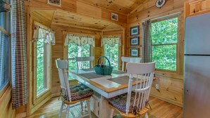 L’intérieur d’un chalet en bois comprend une table à manger, deux chaises, un réfrigérateur et de grandes fenêtres avec des rideaux.