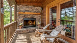 Une terrasse en bois avec une cheminée en pierre, deux fauteuils et une porte vitrée donnant sur une autre terrasse d’où l’on peut admirer la vue sur les arbres.