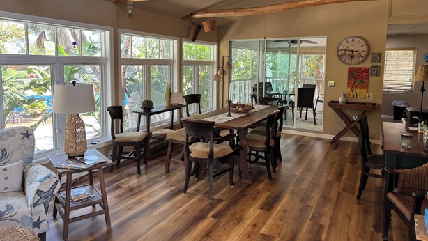 A spacious dining area with a wooden table and chairs, large windows offering a view of greenery, and a ceiling fan.
