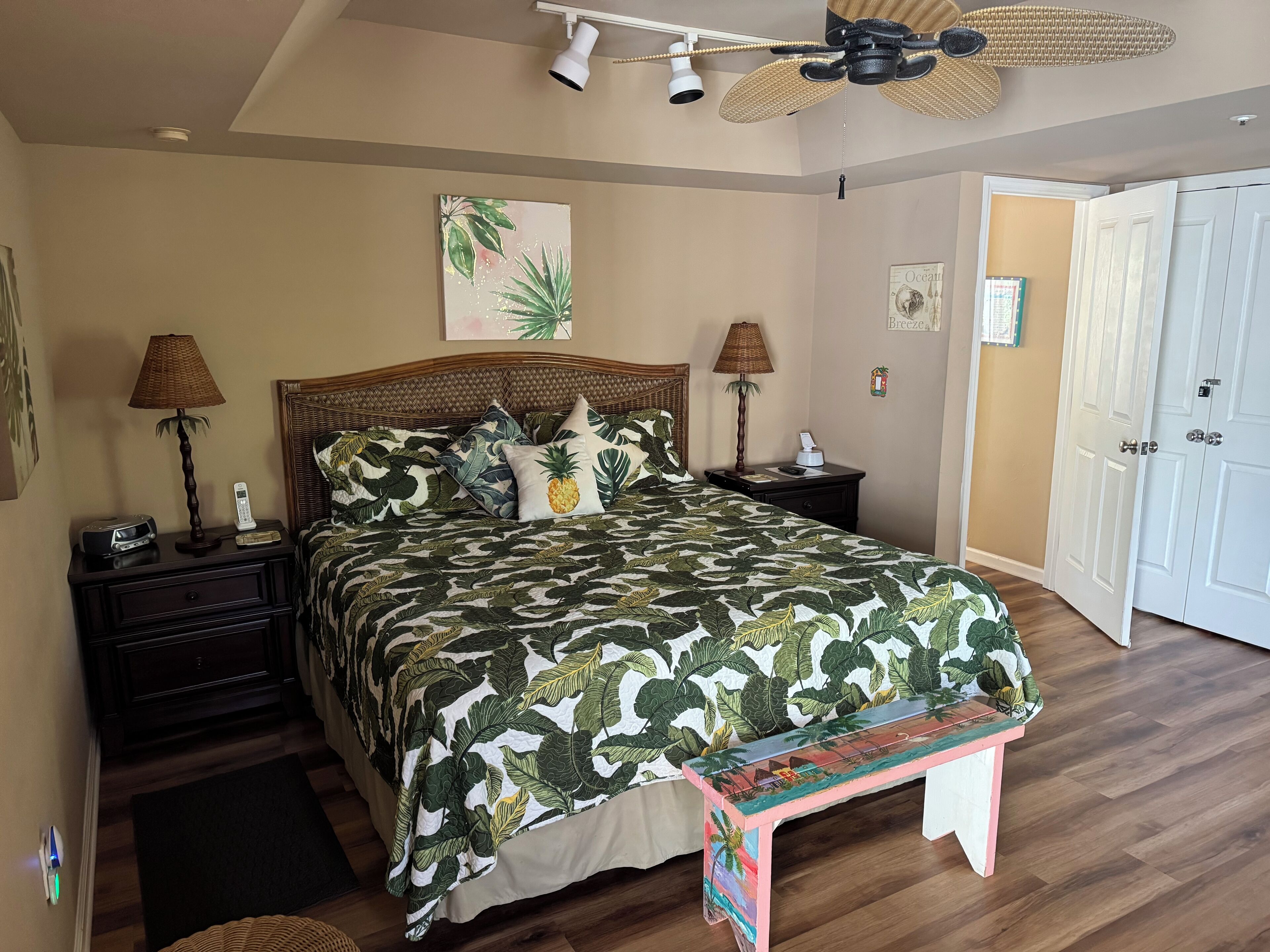 Quarto com cama, mesas de cabeceira, um banco, ventoinha no teto e uma porta.