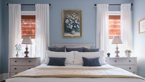 A bedroom with a bed, white curtains, a painting on the wall, and two bedside tables with lamps. - Comfortable Escape 2BR Historic Downtown (Savannah)