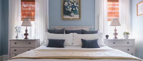 A bedroom with a bed, white curtains, a painting on the wall, and two bedside tables with lamps.