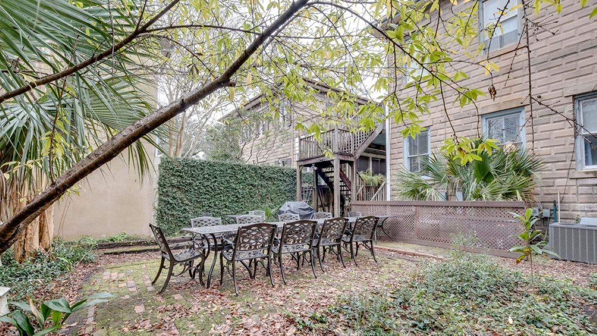 A patio with a table and chairs, surrounded by greenery and a building in the background.