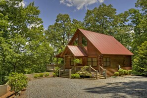 A wooden cabin with a red roof, surrounded by trees and a gravel driveway. - Bear Ridge Hideaway | Hot Tub | Pool Table | MTN View (McCaysville)