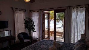 Un dormitorio con cama, una silla, un televisor y un ventanal con cortinas.