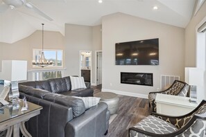 A spacious living room with a leather sofa, a glass coffee table, a fireplace, and a large flat-screen TV mounted on the wall.