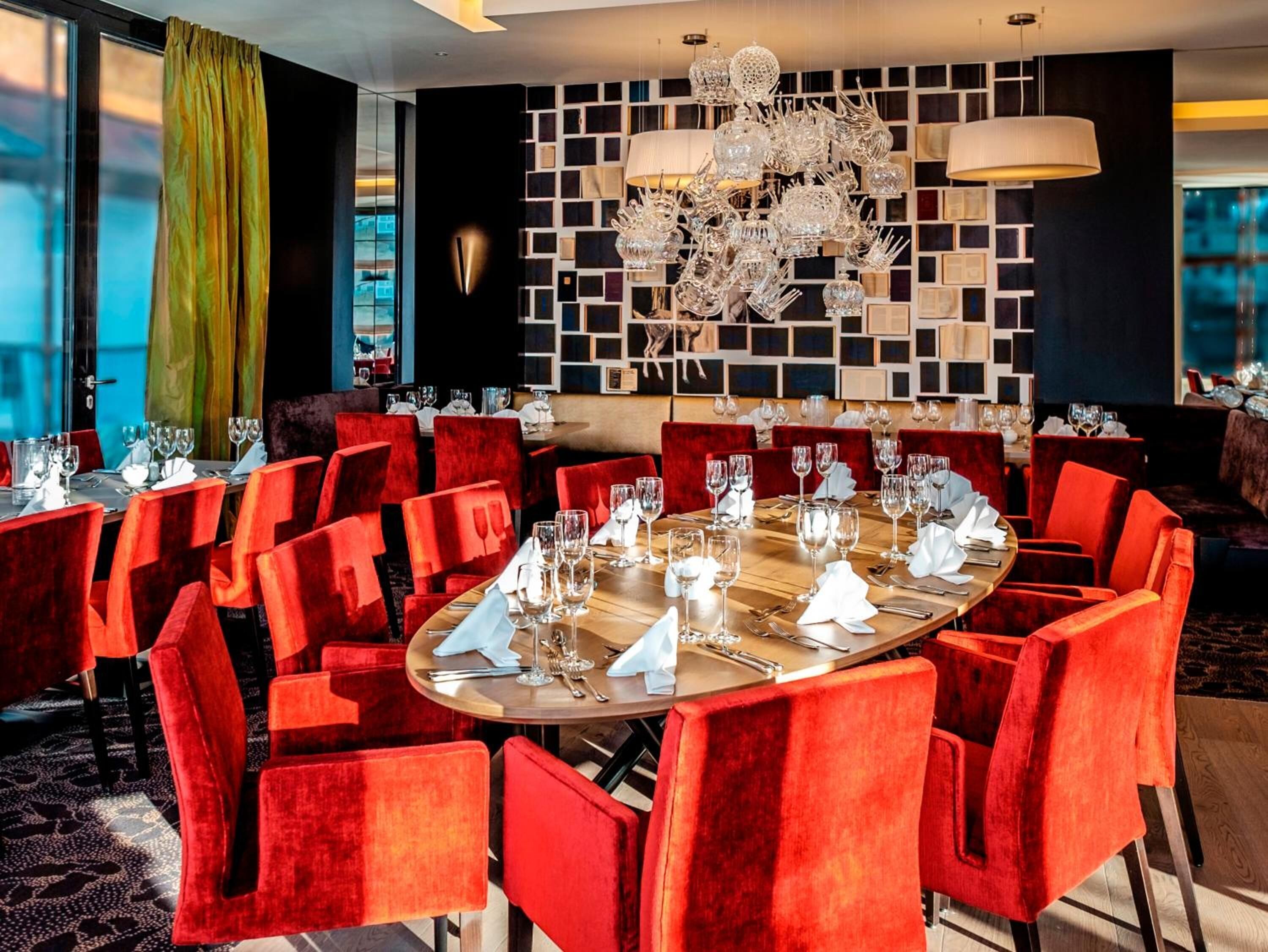 A modern dining area with round tables and red velvet chairs, featuring a decorative wall with framed pictures and a chandelier.