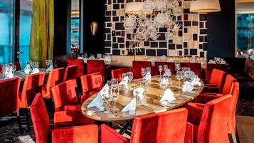 A modern dining area with round tables and red velvet chairs, featuring a decorative wall with framed pictures and a chandelier.