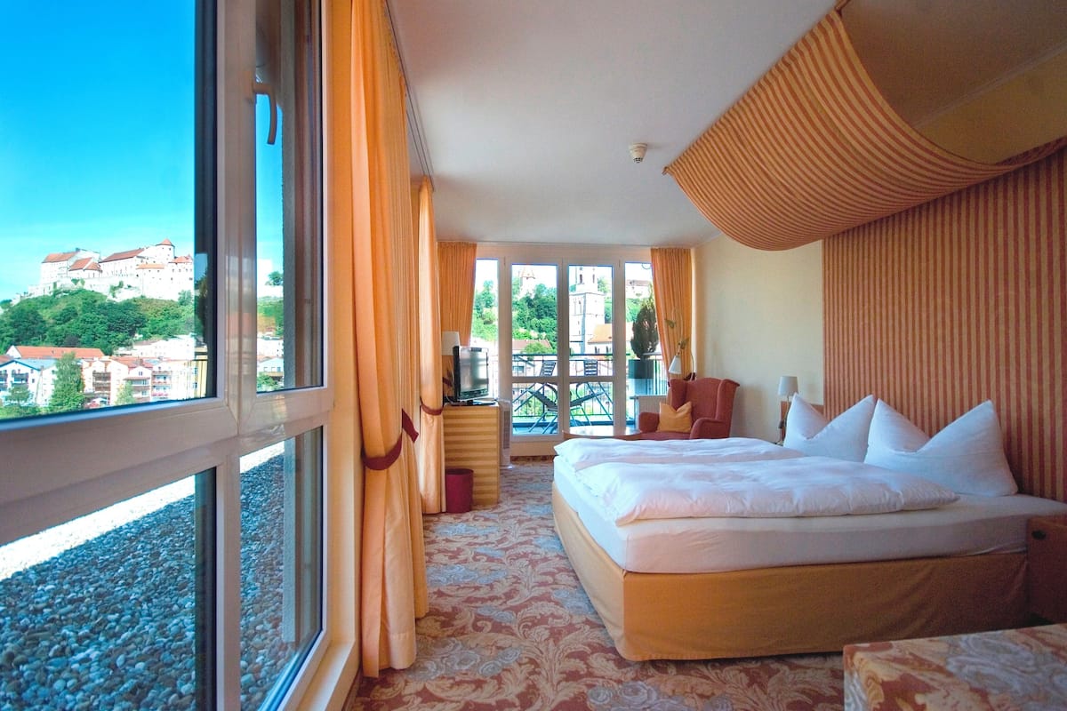 A hotel room with a large bed, a TV, a desk, and a view of a castle through large windows.