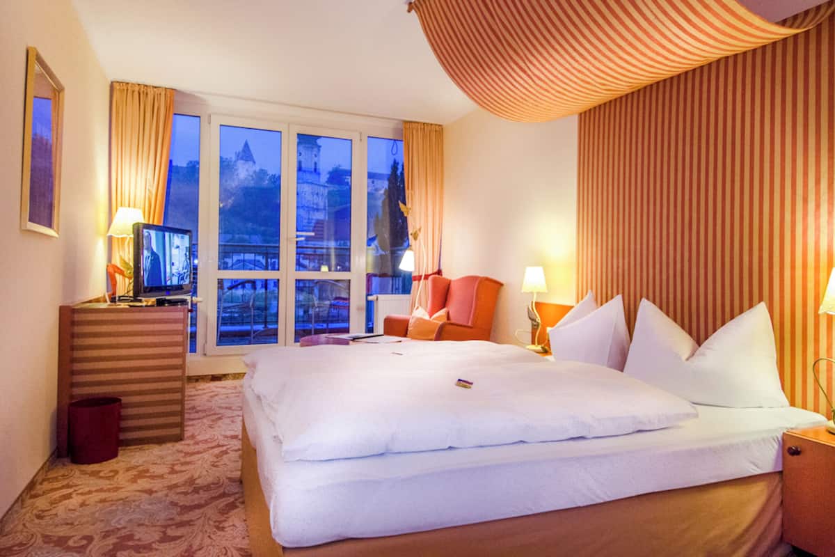 A hotel room with a large bed, a desk with a computer, a chair, a window with curtains, and a view of a building outside.