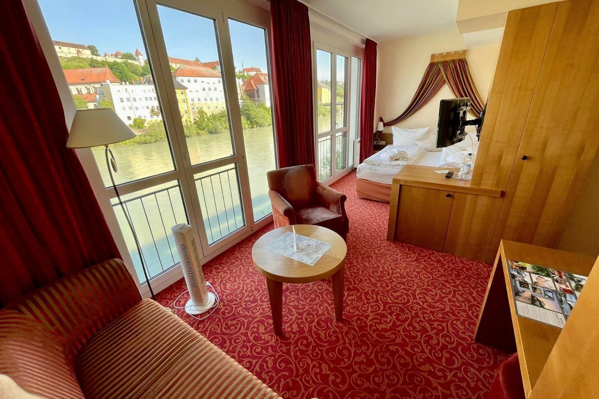 A hotel room with a red carpet, a sofa, a chair, a small table, a bed, and a view of buildings through large windows.