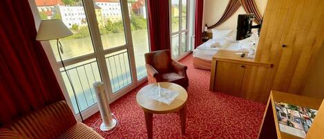 A hotel room with a red carpet, a sofa, a chair, a small table, a bed, and a view of buildings through large windows.