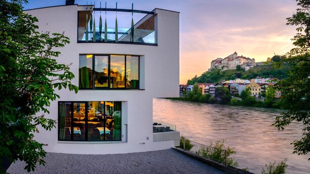 Um edifício moderno com grandes janelas que dão para um rio, com um castelo histórico ao fundo.