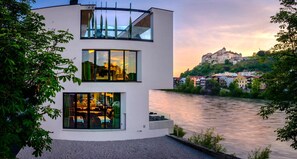 A modern building with large windows overlooking a river, with a historic castle in the background. - Burgblick (Hochburg-Ach)