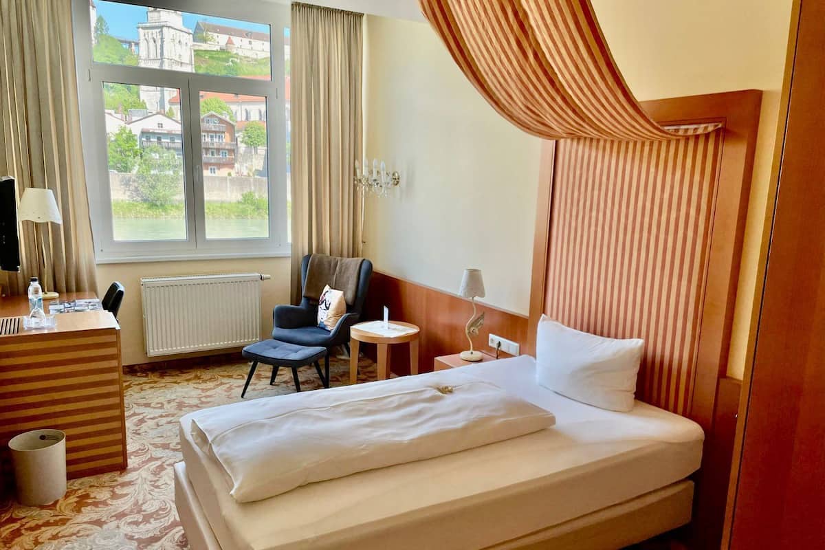 A hotel room with a large bed, a desk with a lamp, a chair, a small table with a lamp, and a view of buildings through the window.