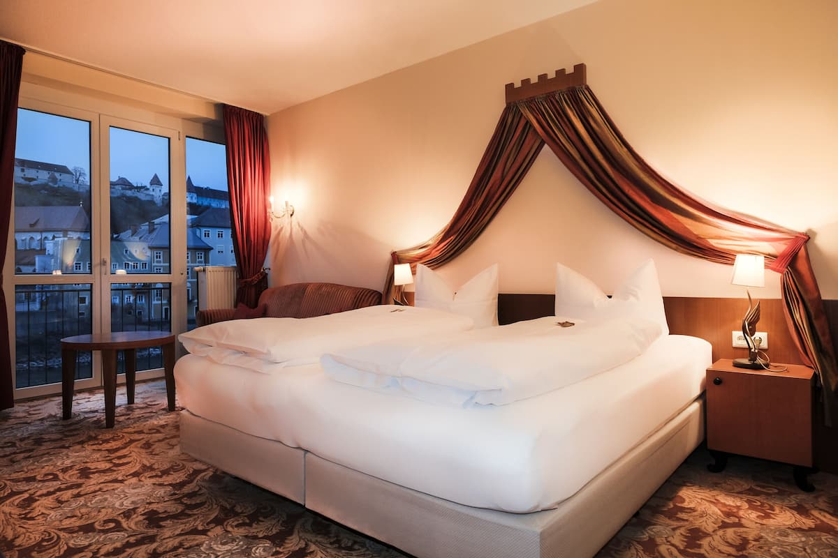 A hotel room with a large bed, a nightstand, a small table, and a view of a cityscape through large windows.
