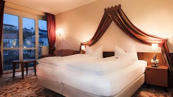 A hotel room with a large bed, a nightstand, a small table, and a view of a cityscape through large windows.