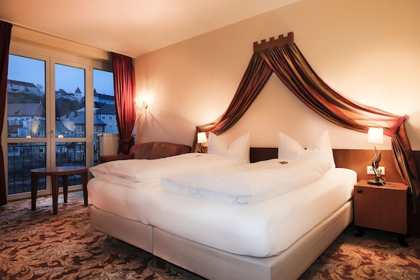 A hotel room with a large bed, a nightstand, a small table, and a view of a cityscape through large windows. - Burgblick (Hochburg-Ach)