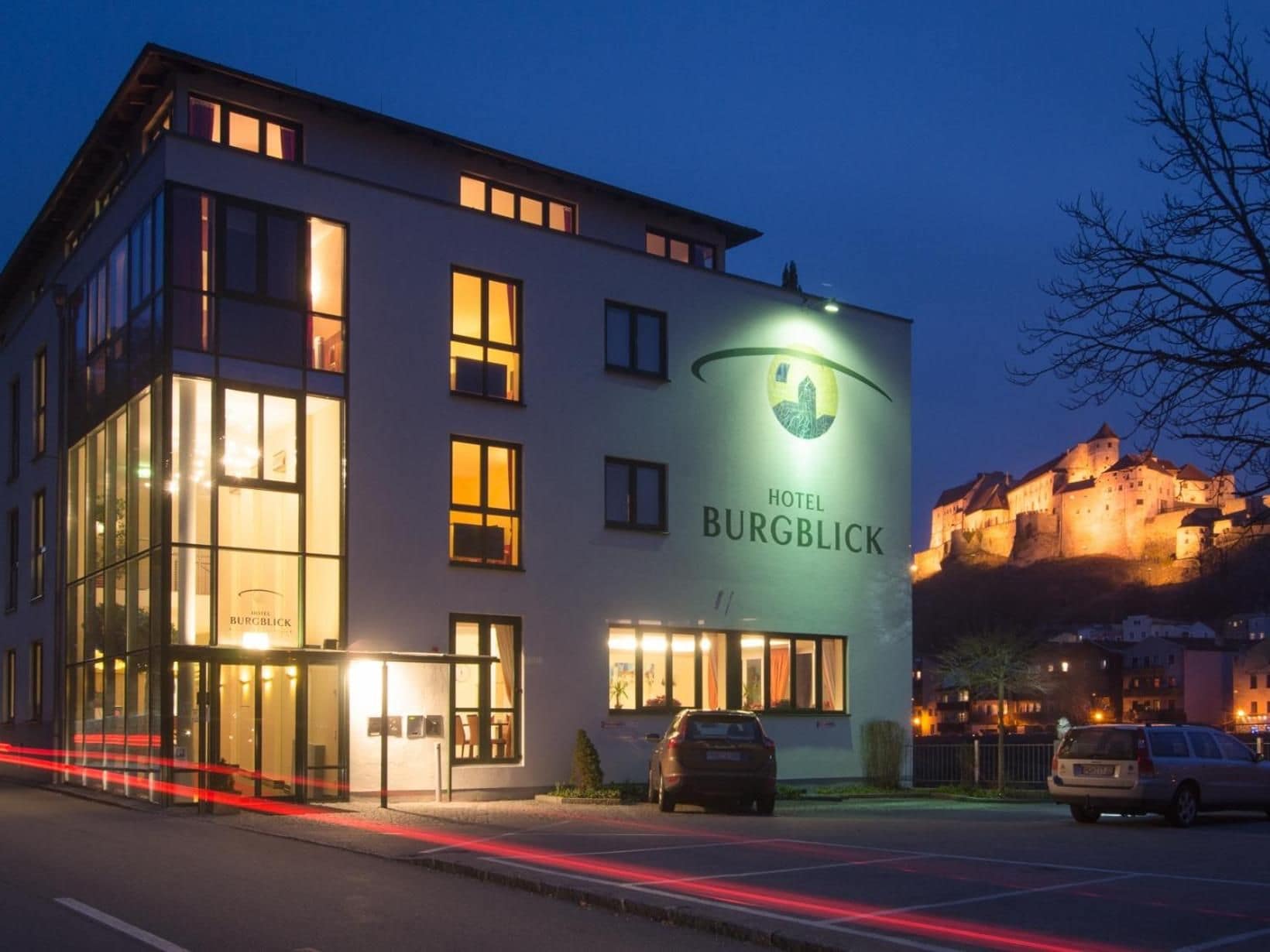 Un bâtiment d'hôtel moderne, avec une enseigne bien visible indiquant « HOTEL BURGBLICK », de grandes fenêtres et un parking où sont garées des voitures.