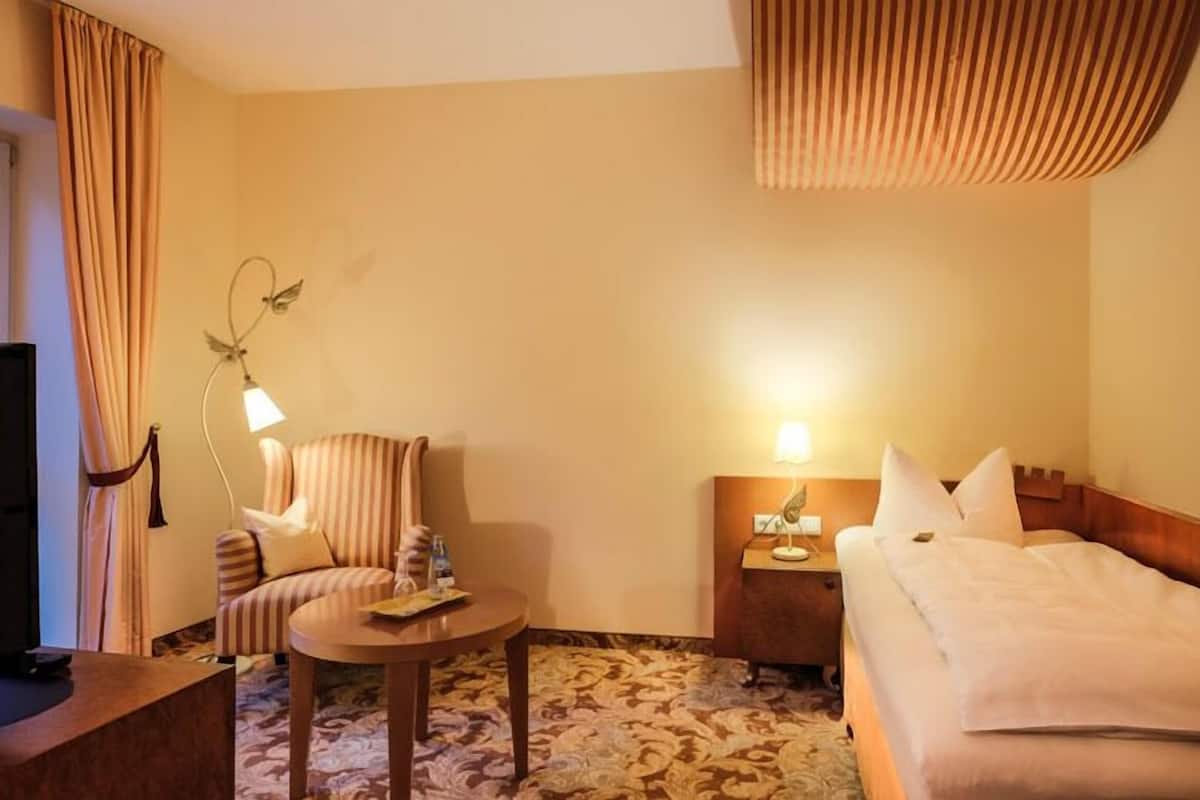 A hotel room with a bed, a chair, a small table, a nightstand, and a lamp.