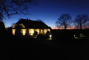 A house with a dark exterior and illuminated windows, surrounded by trees and a clear sky. - Gut Rattelvitz (Gingst)