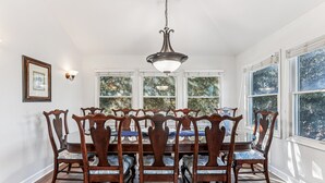 A dining room with a wooden table and chairs, large windows, and a hanging light fixture.