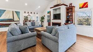 A living room with two grey sofas, a wooden coffee table, and a TV on a wooden stand. There is a bookshelf with various items and a painting of a sailboat on the wall.
