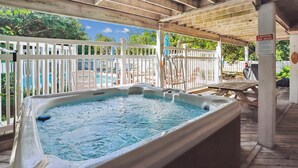 A hot tub under a wooden pergola with a white fence, a pool in the background, and a sign indicating spa rules.