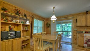 A wooden cabin interior with a dining table, chairs, a kitchen area, and a large window with blinds. - Toccoa River Escape | River Front | Gas Grill (Mineral Bluff)