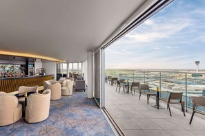 A modern lounge area with a bar, comfortable seating, and a view of an airport. - Hilton Garden Inn London Heathrow Terminal 2 and 3 (Hounslow)