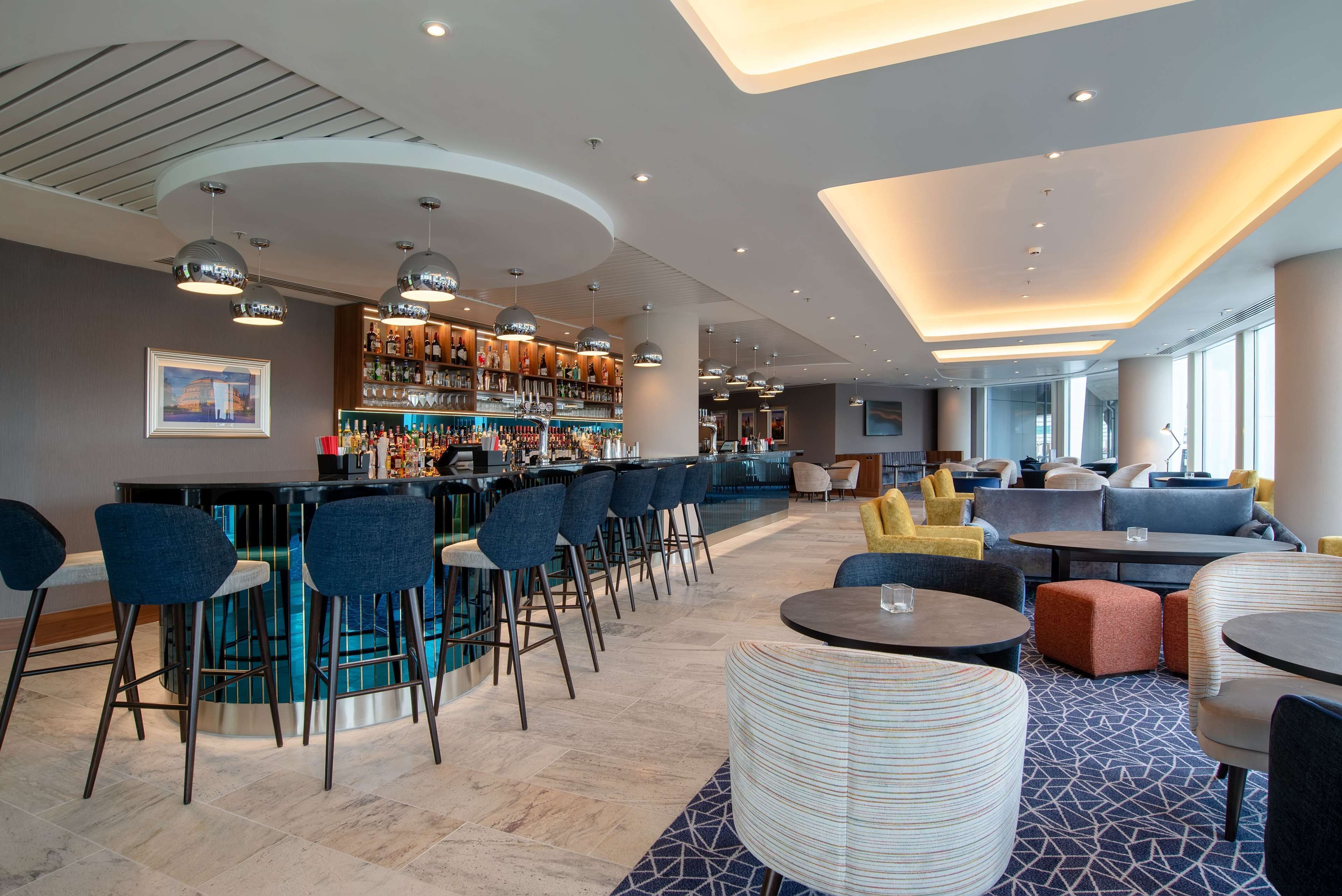 A modern bar area with a curved counter, multiple seating options including bar stools and round tables, and a patterned carpet.