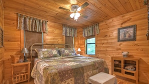 A cozy wooden cabin bedroom with a bed, a nightstand, a lamp, a window with curtains, a ceiling fan, and a small cabinet.
