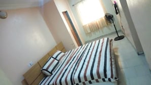 Un dormitorio con una cama, una ventana con cortinas, un ventilador de pie y un enchufe de pared.