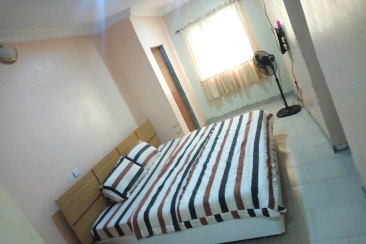 Un dormitorio con una cama, una ventana con cortinas, un ventilador de pie y un enchufe de pared.