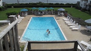Una piscina rectangular con una terraza de madera alrededor, vista al océano, y varias sillas de descanso y sombrillas.