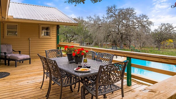 Una terraza de madera con una mesa y sillas, una casita con techo de metal y una piscina al fondo.