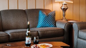 Una sala de estar acogedora con un panel de madera, una obra de arte enmarcada, un sofá con un cojín azul, una mesa de centro con una botella y vasos, y una lámpara de mesa.