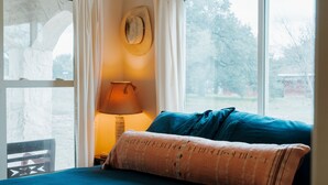 Una habitación con una cama, una lámpara, un sombrero en la pared y vistas a árboles por una ventana.