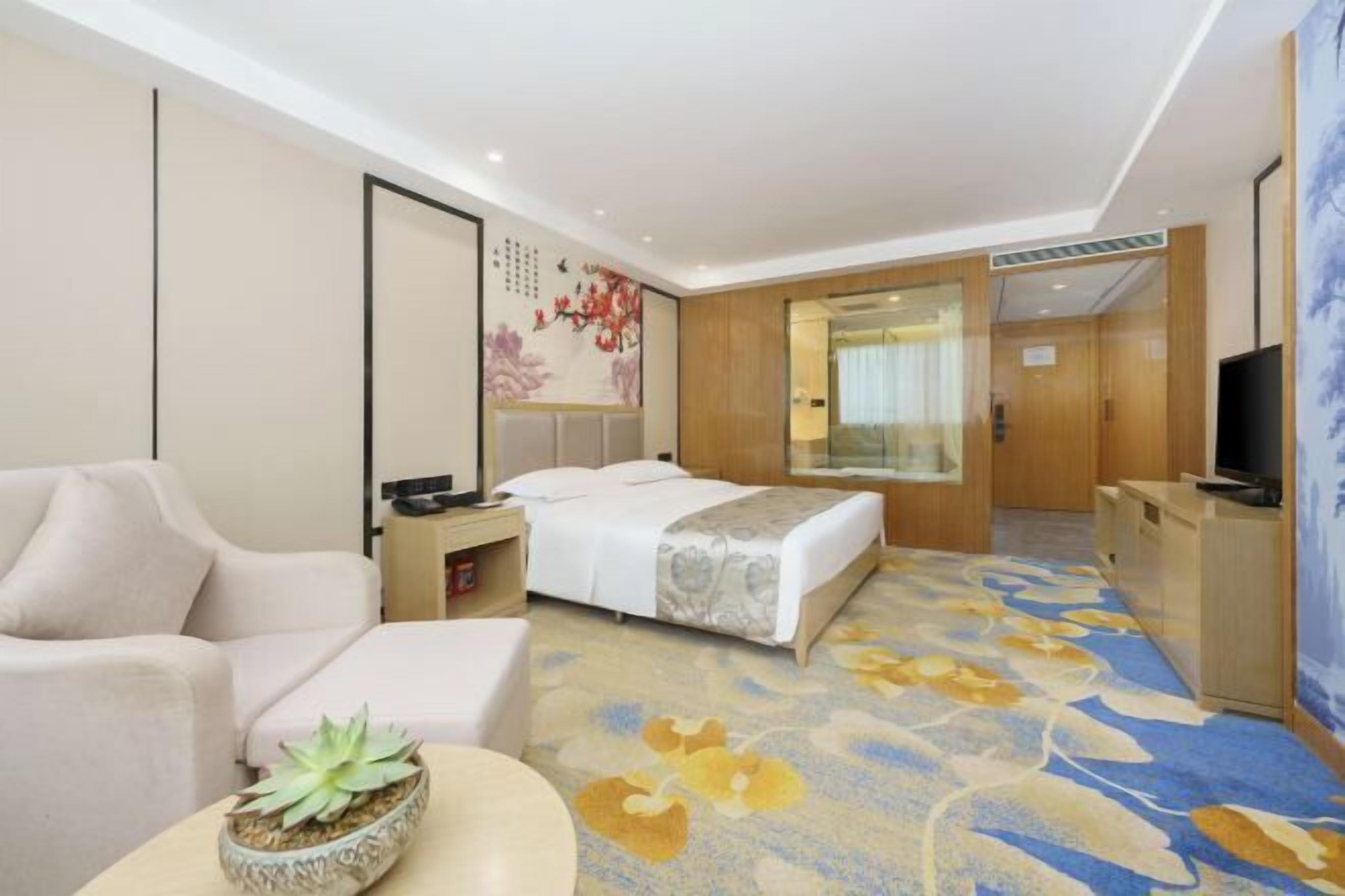 Une chambre d’hôtel avec un lit, un canapé, une petite table sur laquelle est posée une plante, un téléviseur et une moquette à motifs floraux.