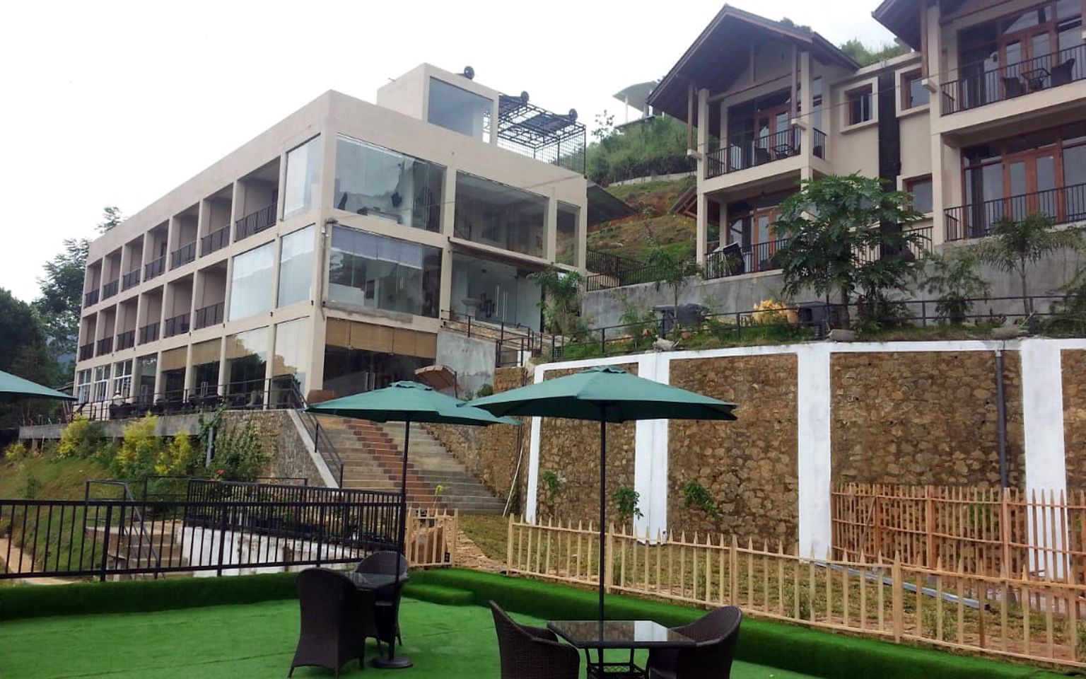 A modern hotel with multiple floors, outdoor seating with umbrellas, and a stone retaining wall.