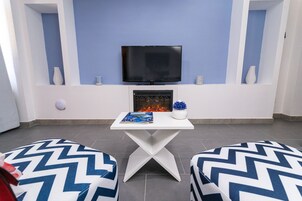 A modern living room with a flat-screen TV mounted above a fireplace, two patterned ottomans, and a small table with a book and vase.